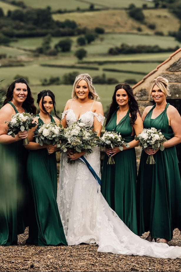 A bride in a white lace wedding gown stands with four bridesmaids wearing matching dark green sleeveless dresses, holding white and green floral bouquets outdoors in front of a stone building and rolling green hills.